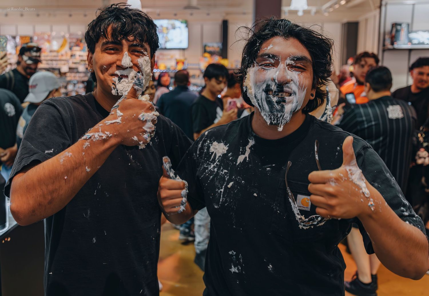 Two young men smiling with cake on their faces giving thumbs up.