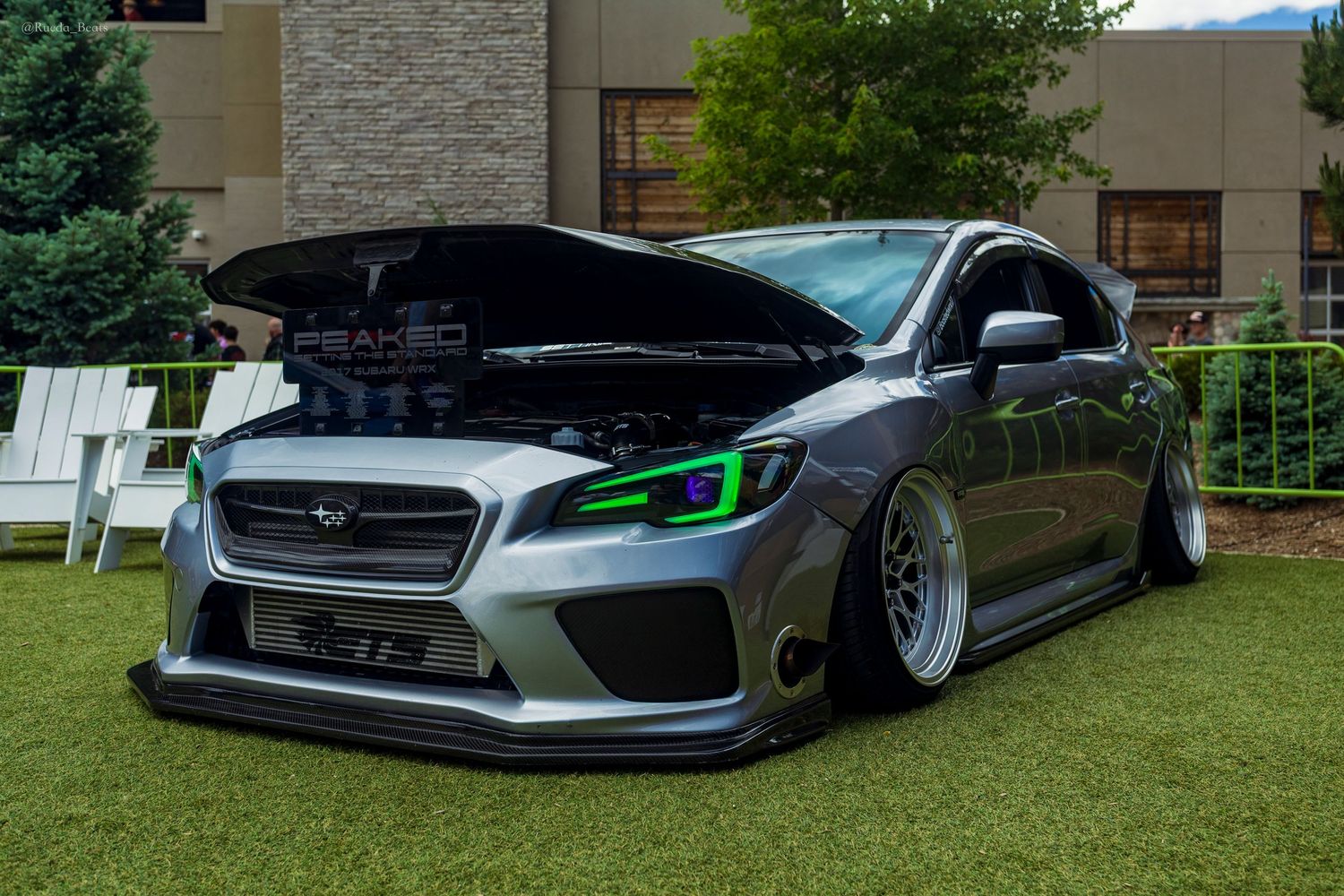 Modified grey Subaru WRX STI with green headlights and open hood displayed at an outdoor car show.