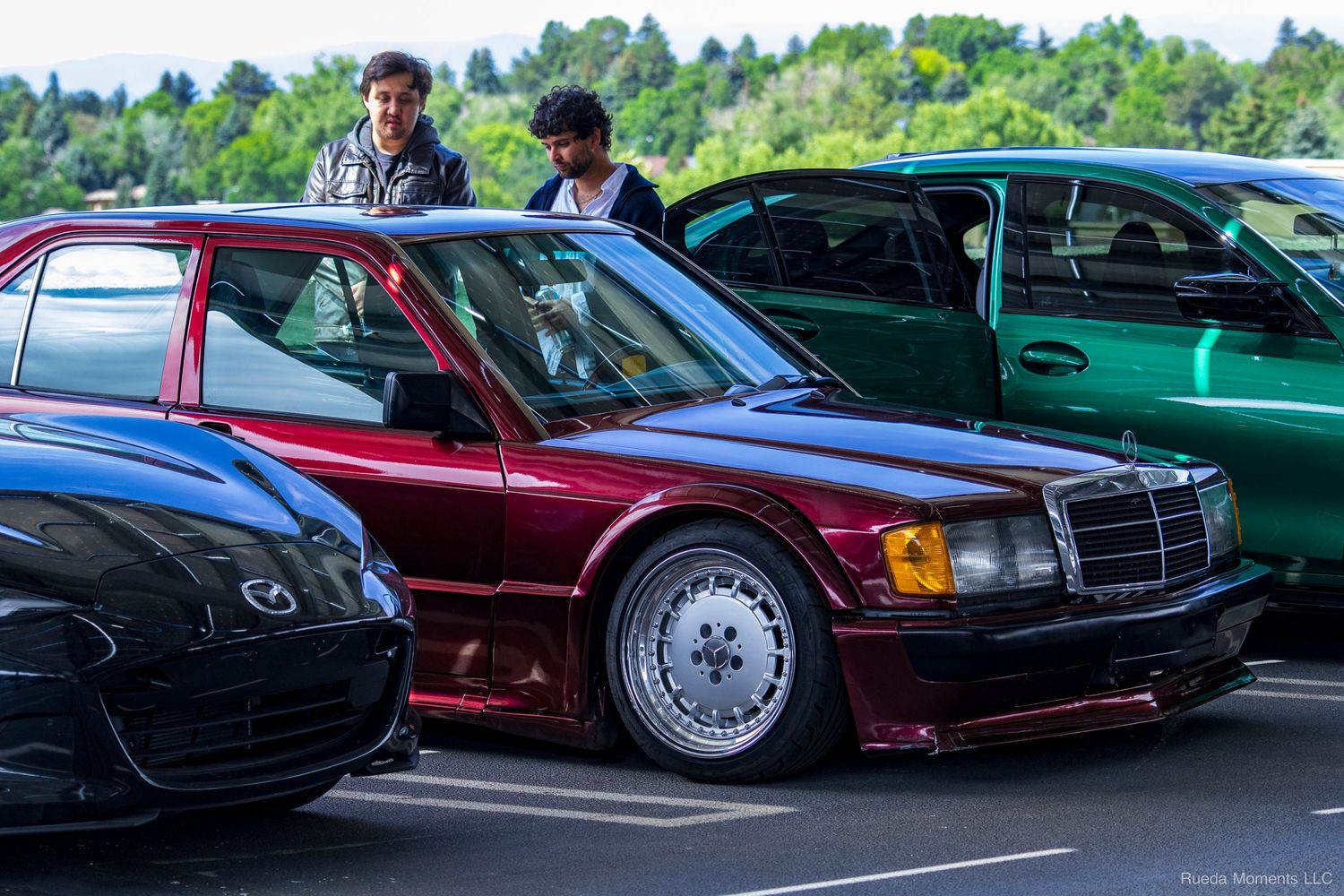 A burgundy Mercedes-Benz 190E with wide body kit parked at a car show between other vehicles.