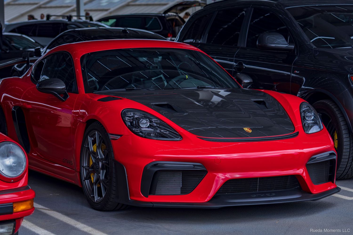 Red Porsche 718 Cayman GT4 with carbon hood parked among other sports cars at a meet.