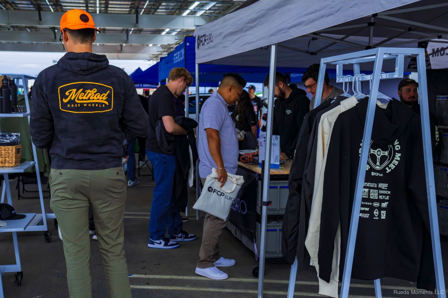 Car enthusiasts browsing merchandise vendor booth at an outdoor automotive event.