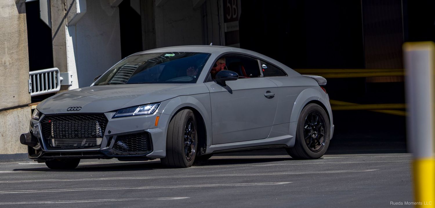 Matte gray Audi TT RS coupe driving through a parking structure at a car event.
