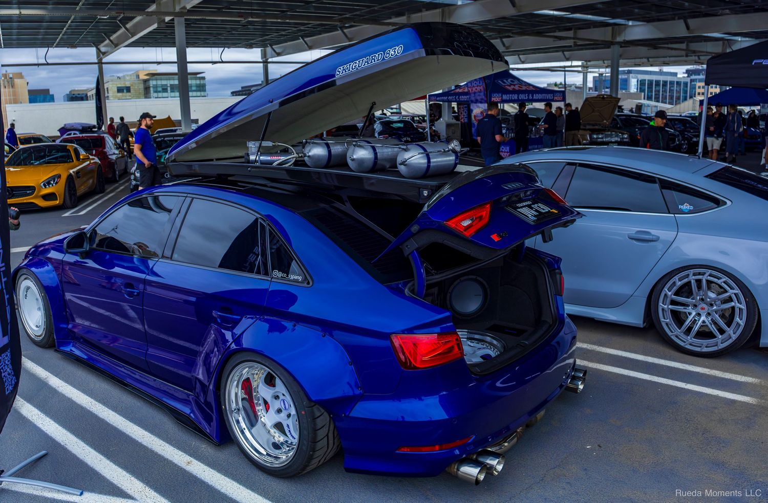 Modified blue Audi RS3 sedan with open trunk and roof rack on display at a car show.