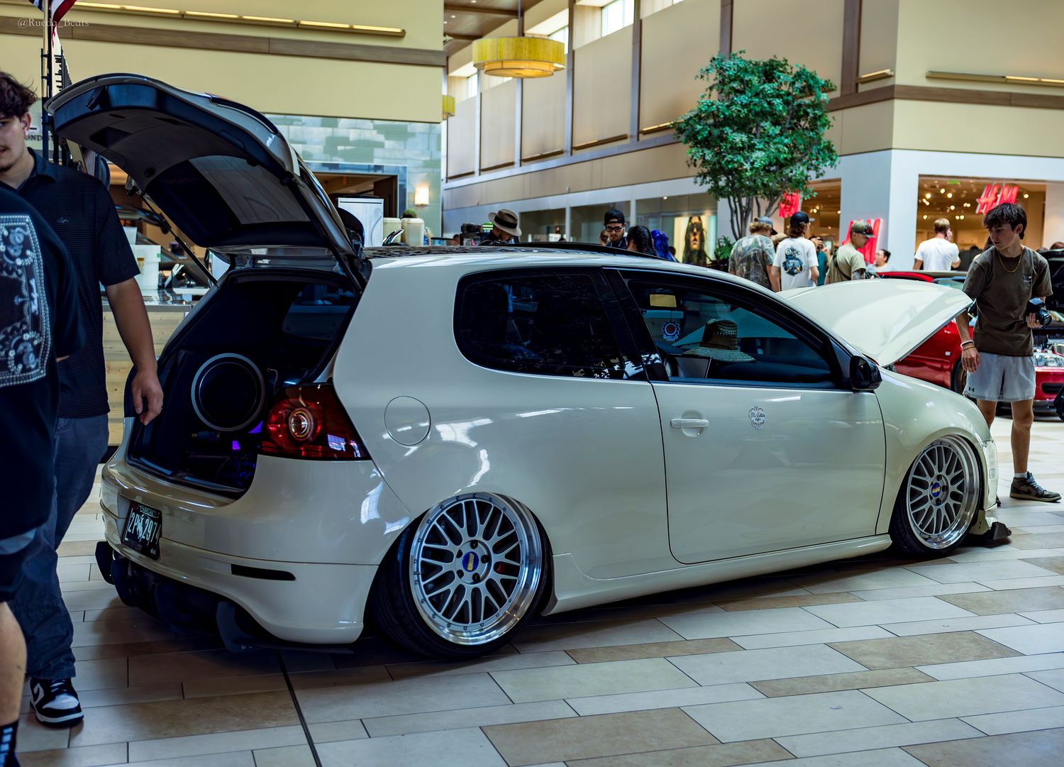 White slammed Volkswagen Golf MK6 with open hatch and subwoofer displayed inside a shopping mall.