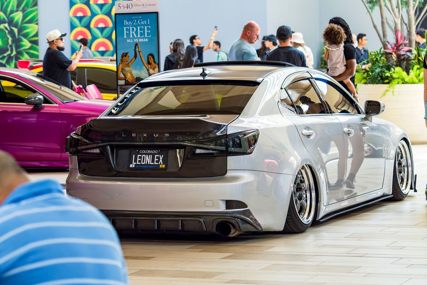 Slammed silver Lexus IS with LEONLEX license plate and aggressive stance at indoor mall car show.