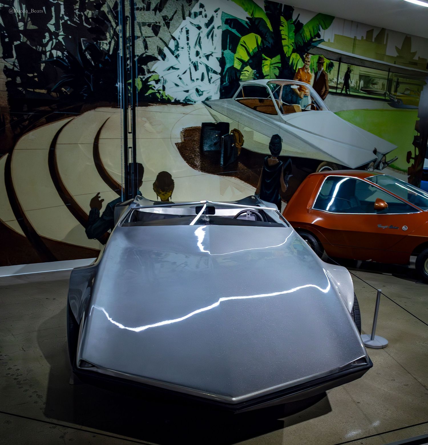 Futuristic angular silver concept car on display in a museum with colorful mural backdrop.