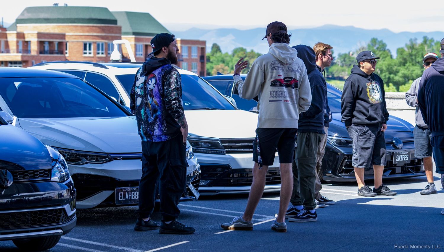 Group of car enthusiasts gathered in a sunny parking lot admiring modified vehicles at an outdoor car meet.
