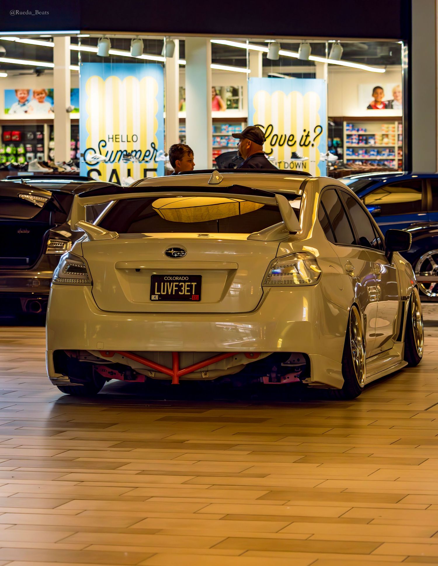 Modified white Subaru WRX with large rear spoiler and custom license plate LUVF3ET displayed at indoor car show.