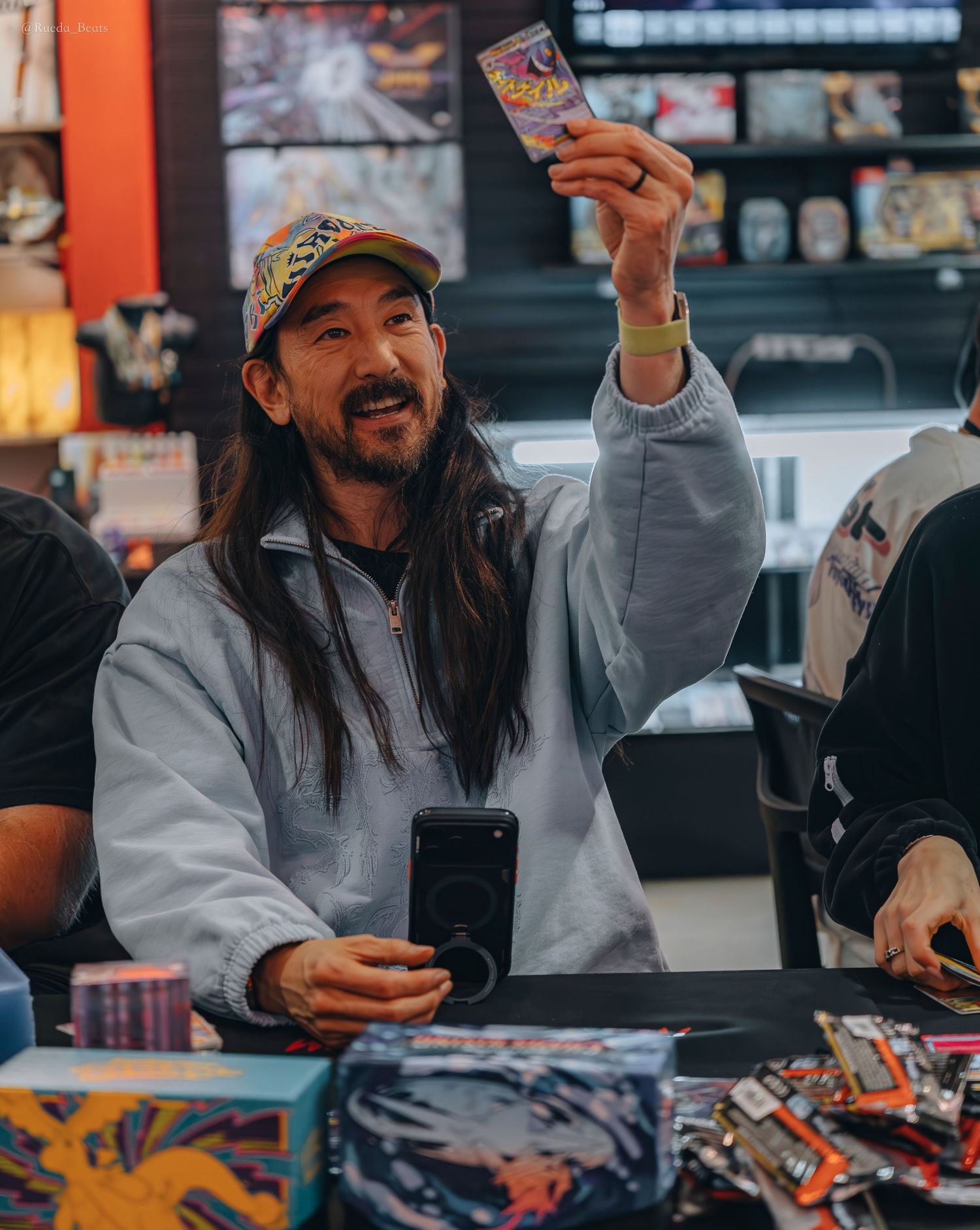 Smiling long-haired man holding up a Pokemon card triumphantly at a trading card opening event.