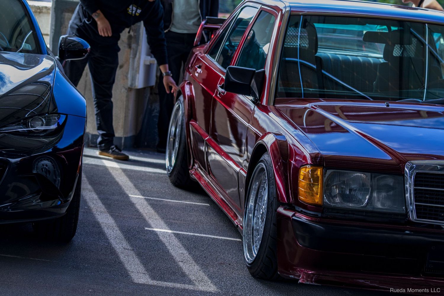 Close-up of a lowered burgundy Mercedes-Benz 190E with wide fenders at a car meet.