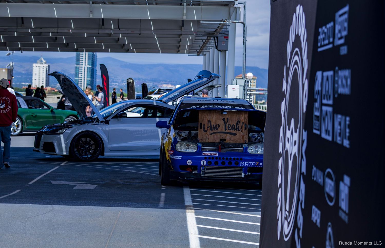 Two modified cars including an Audi R8 at an outdoor car show with event banners.