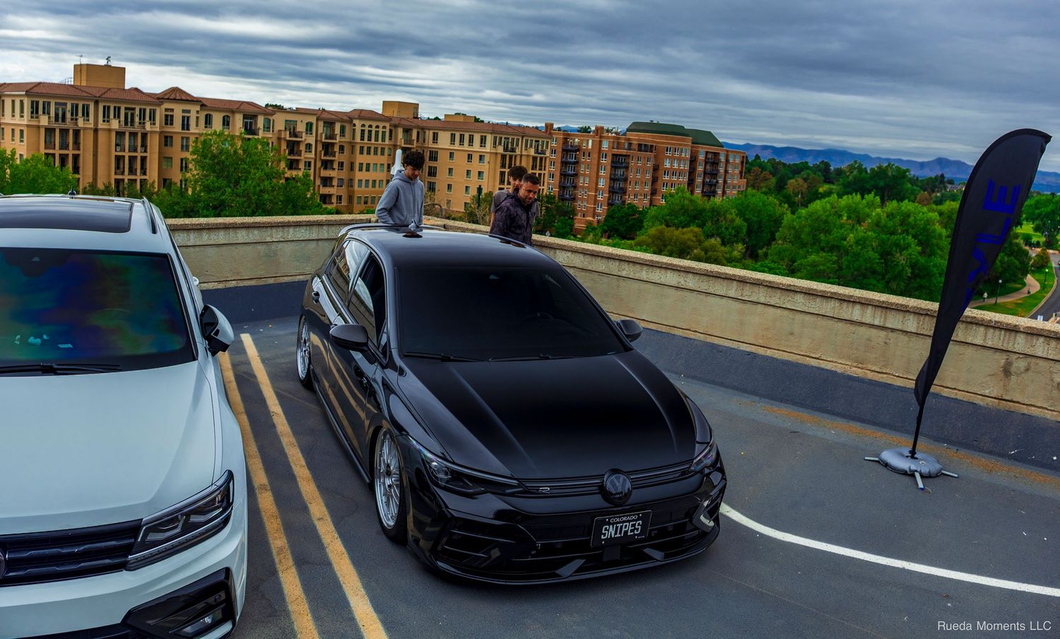 Lowered black Volkswagen Jetta on display at a rooftop car meet with city buildings in background.