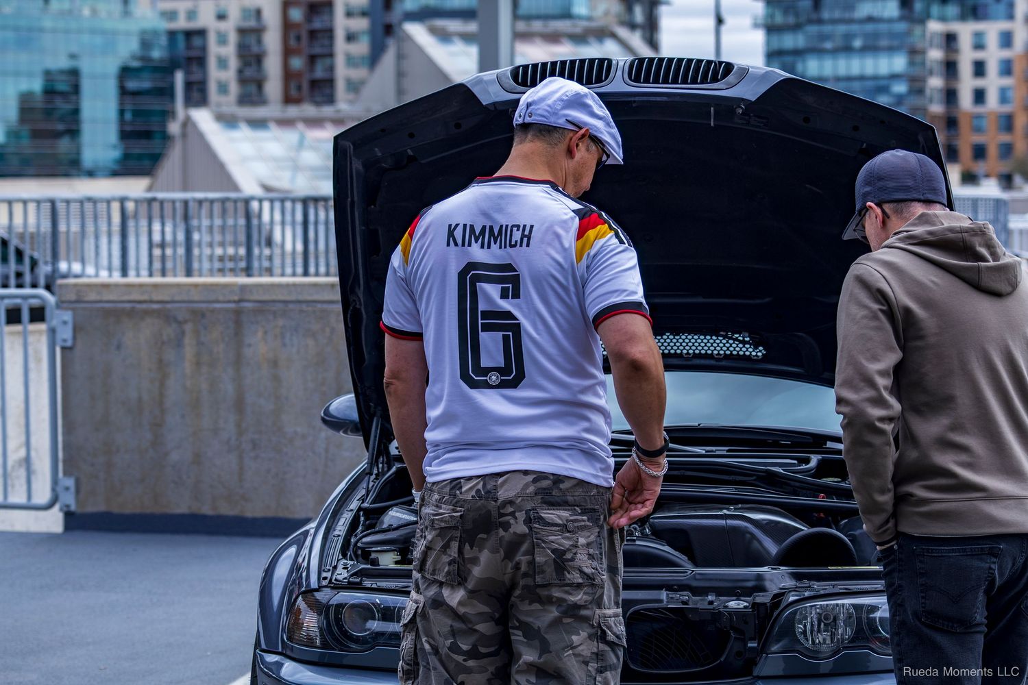 Man wearing Germany jersey number 6 inspecting engine bay of a black car at a meet.