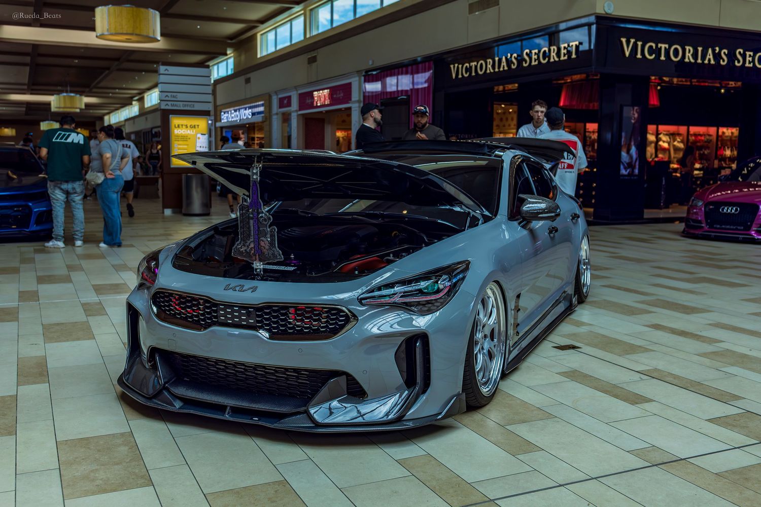 A widebody Kia Stinger GT in gray with aggressive aero kit displayed near Victoria's Secret in a mall.
