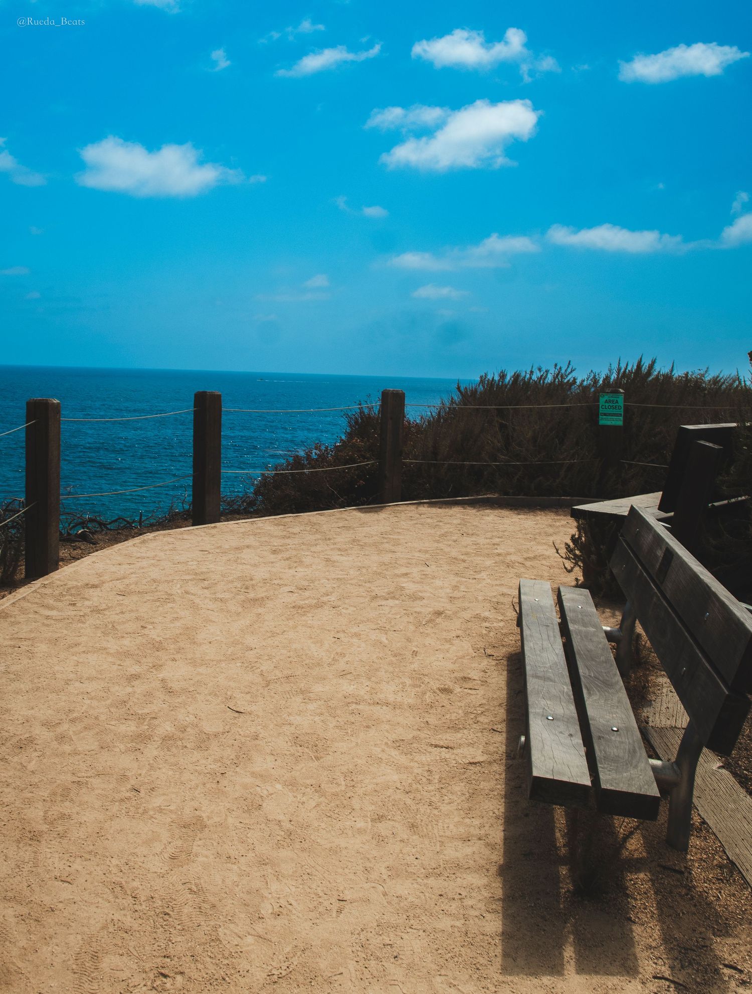 A wooden bench overlooks a sandy coastal overlook with ocean views under a bright blue sky.