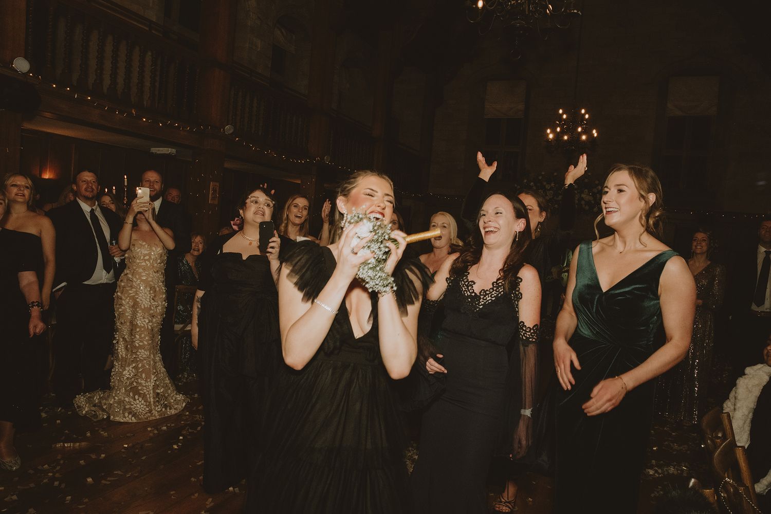 A bridesmaid catches the bouquet toss at an elegant wedding reception, surrounded by guests in formal attire.