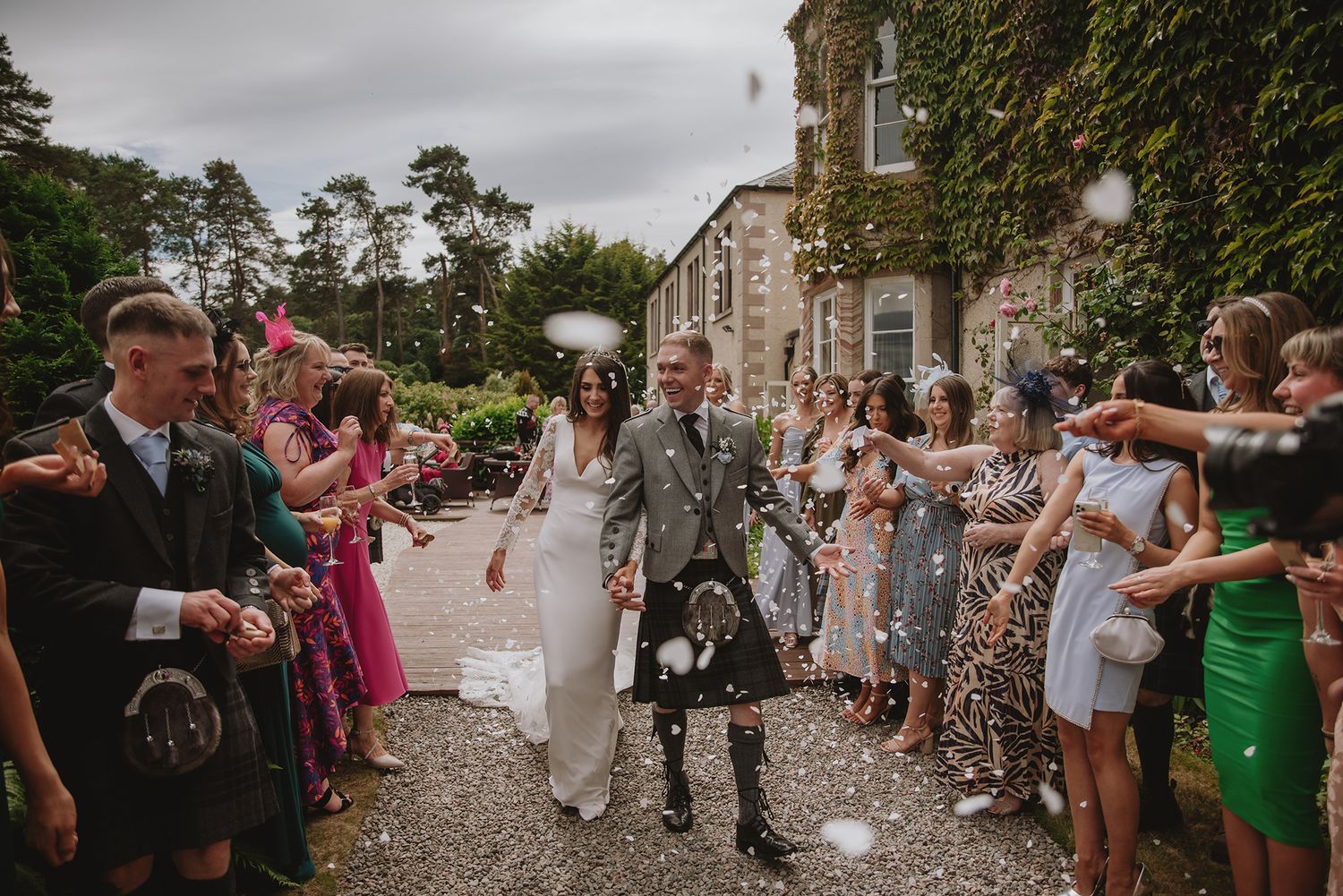 Bride and groom in kilt walk through confetti shower outside ivy-covered Scottish wedding venue with guests cheering.