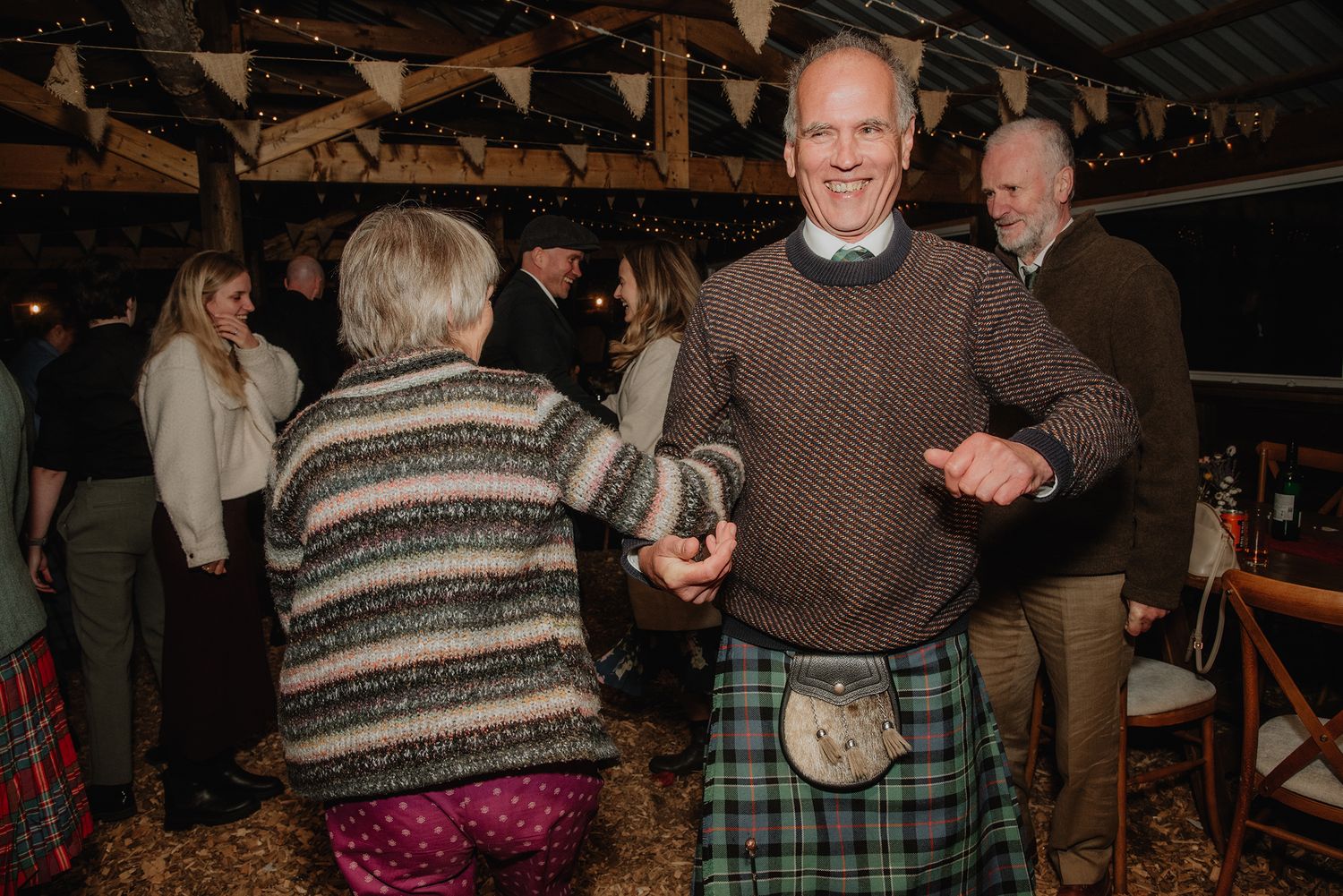 Man in a tartan kilt dancing with a partner at a Scottish wedding reception in a rustic barn venue.