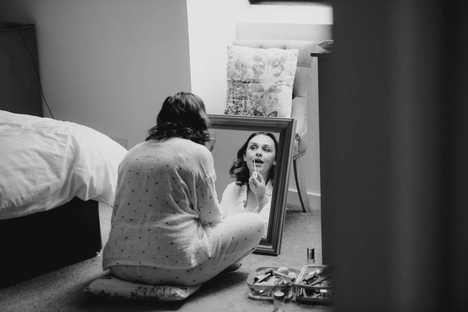 Bride applies lipstick while gazing into a floor mirror in a softly lit bedroom.