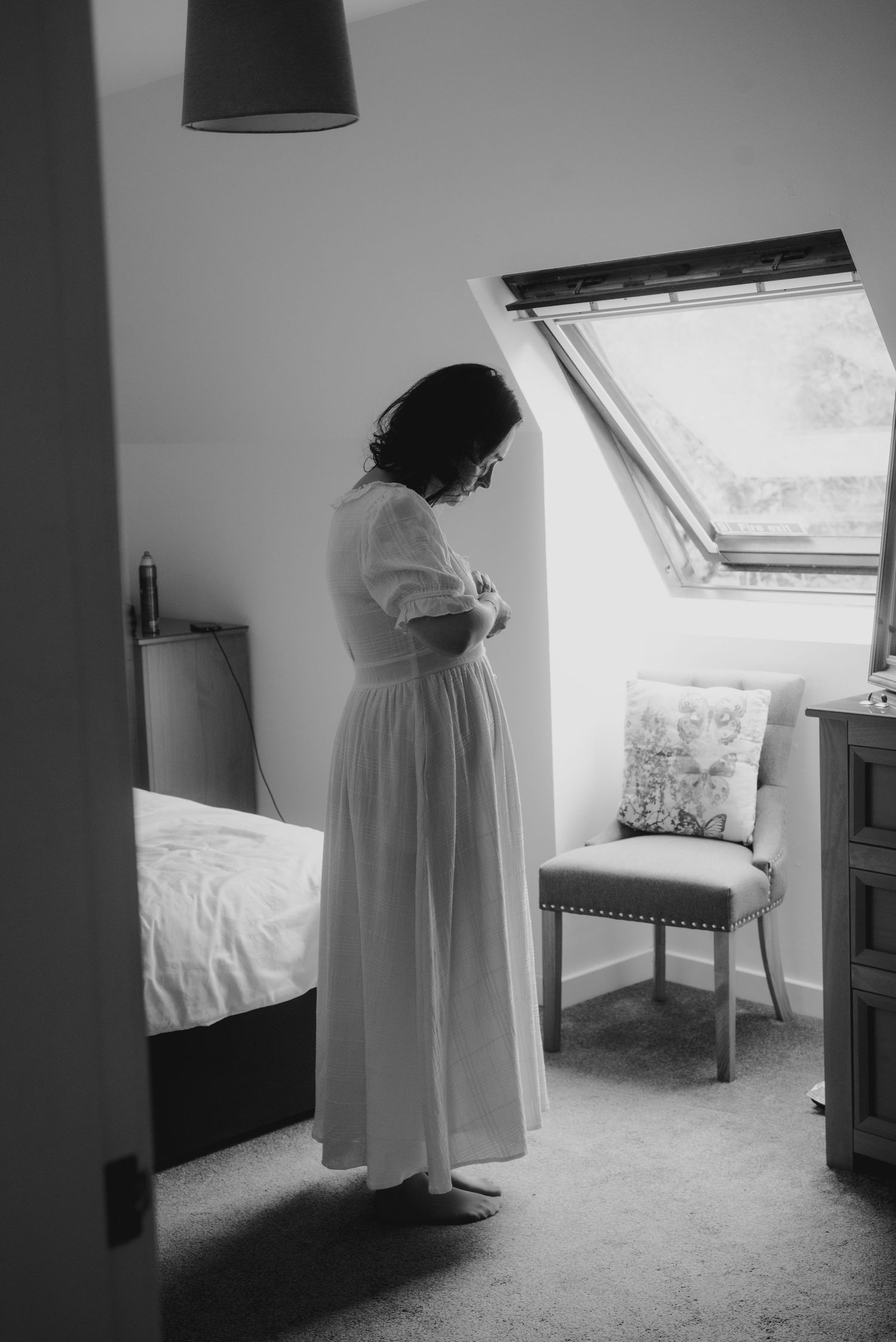 Bride in elegant white wedding dress stands in sunlit attic bedroom adjusting her gown.