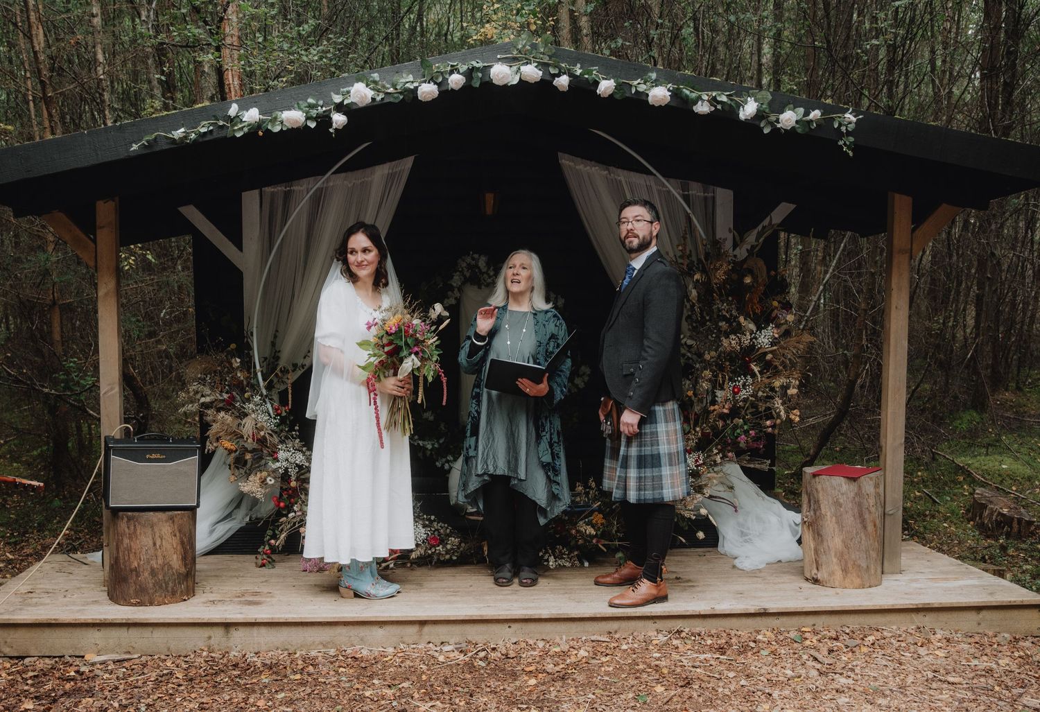 Bride and groom pose with officiant at rustic woodland wedding ceremony shelter with floral decor.