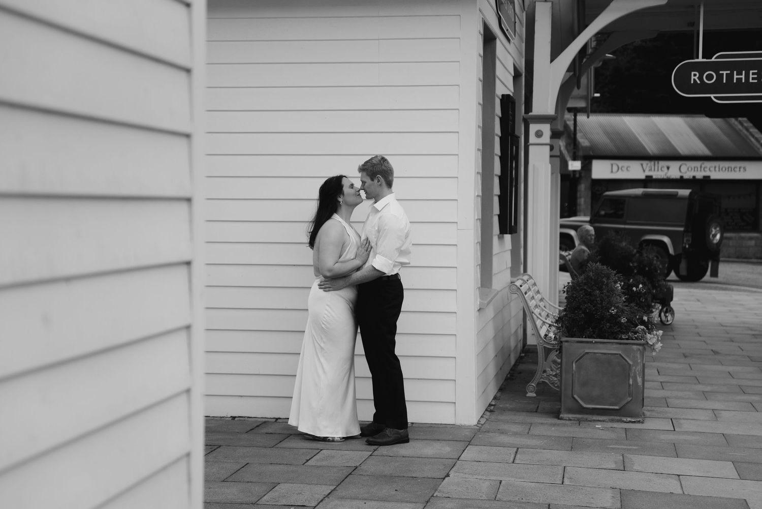Black and white photo of a couple sharing a romantic moment outside a historic building on a cobblestone street.