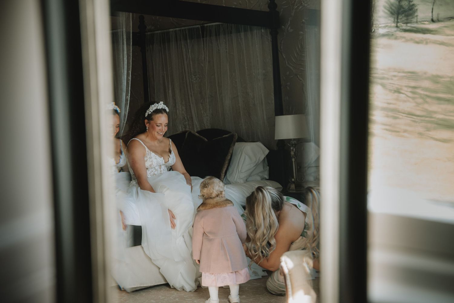 Bride in white gown wearing tiara sits on bed while flower girl and bridesmaid help with final wedding preparations.