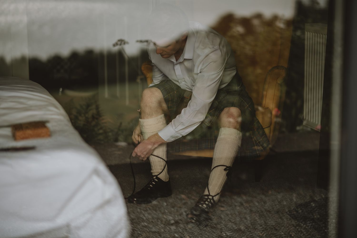 Groom in Scottish kilt lacing traditional ghillie brogues shoes, reflected through rain-streaked window.