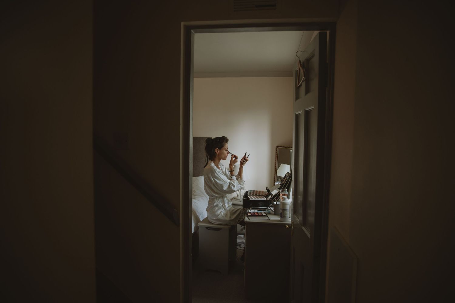 Bride in white robe applying makeup at a vanity table before the wedding.