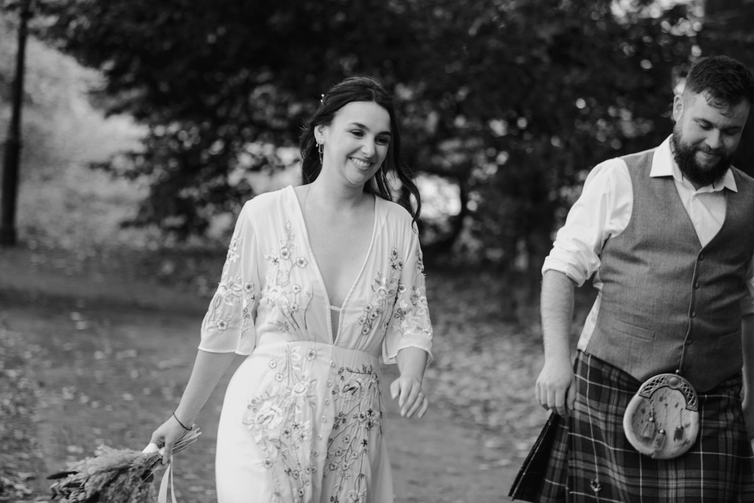 Black and white photo of smiling bride in floral dress walking with kilted groom at outdoor wedding.