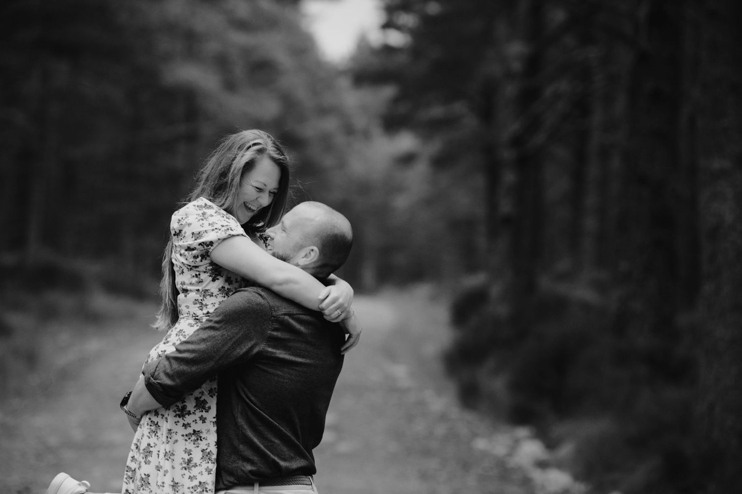 Romantic black and white photo of couple embracing on a forest path surrounded by tall pine trees.