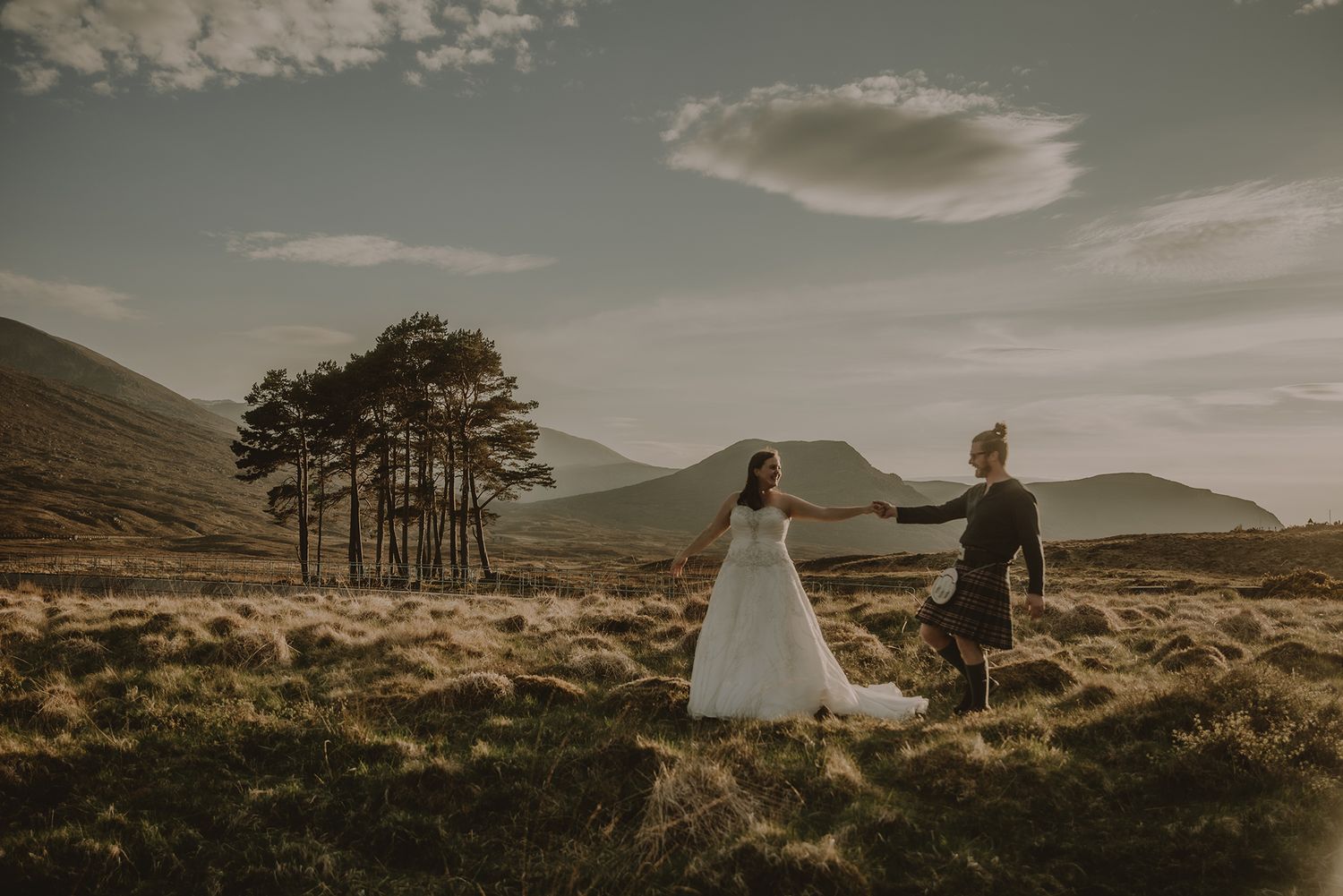 Bride in a white gown and groom in a Scottish kilt holding hands in a golden highland landscape with pine trees.