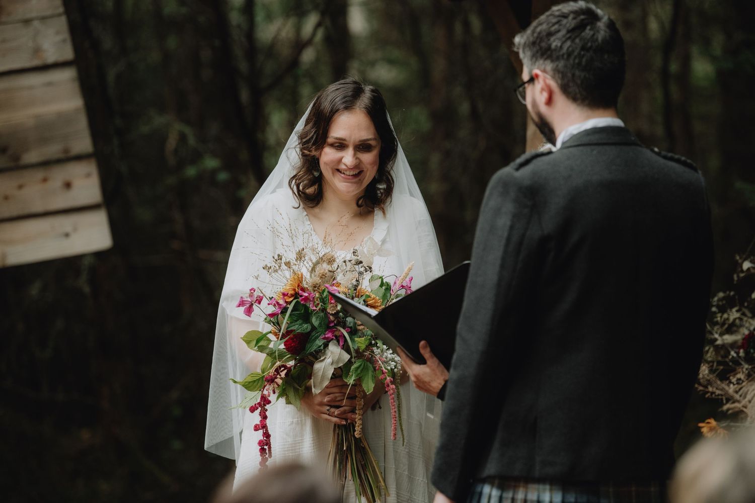 Smiling bride in white gown and veil holds colorful bouquet during intimate woodland wedding ceremony.