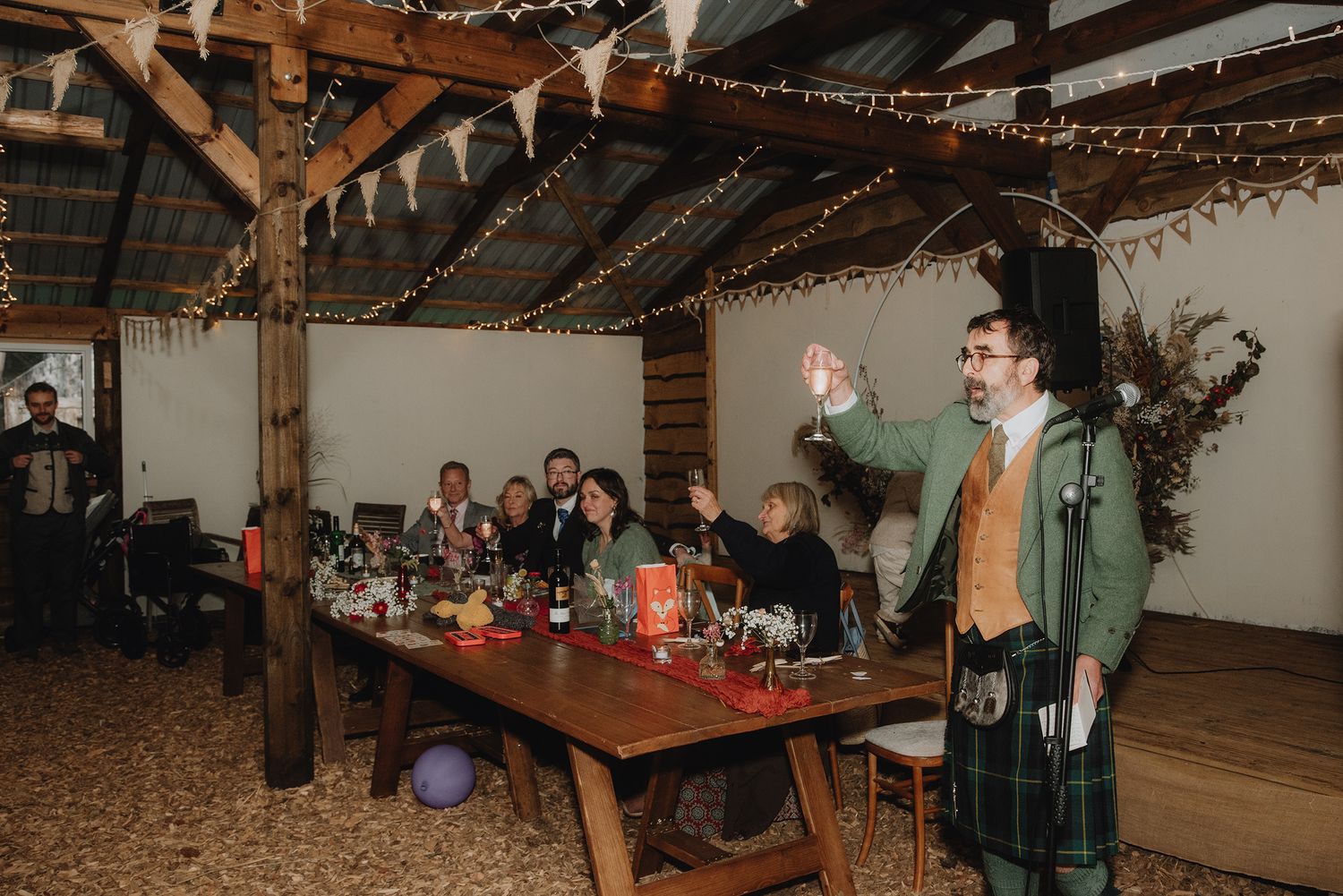 Kilted wedding speaker raising hand while addressing guests at decorated rustic barn reception.