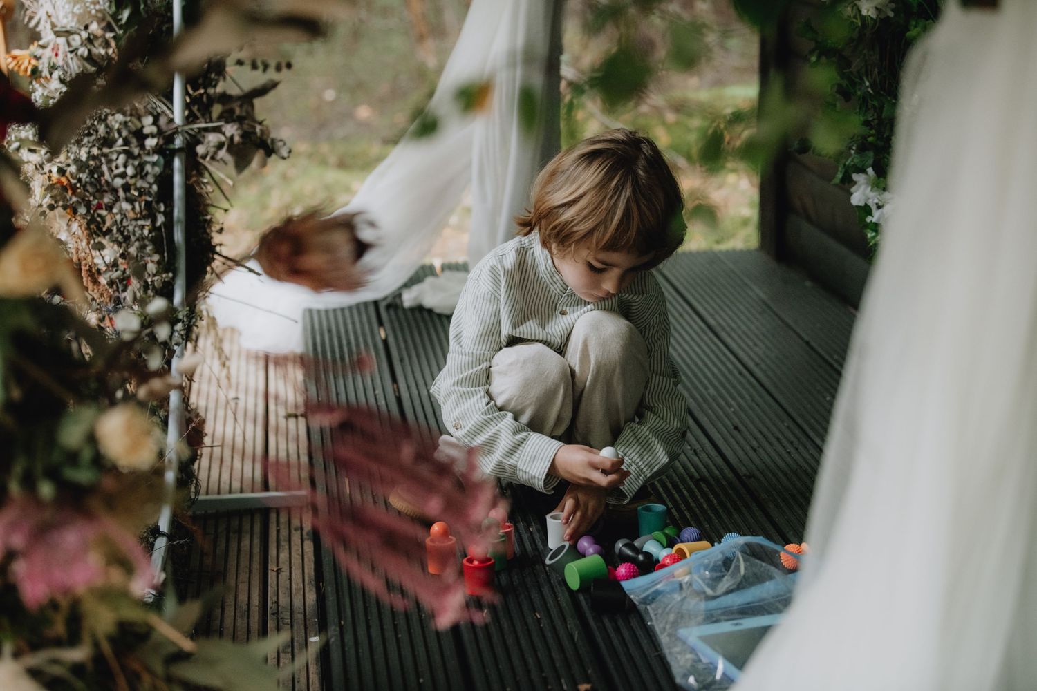 Young child plays with colorful toys on wooden deck during outdoor woodland wedding ceremony.