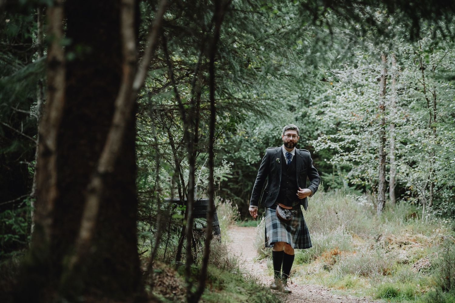 Man in traditional Scottish kilt and jacket walking along a forest path surrounded by tall pine trees.