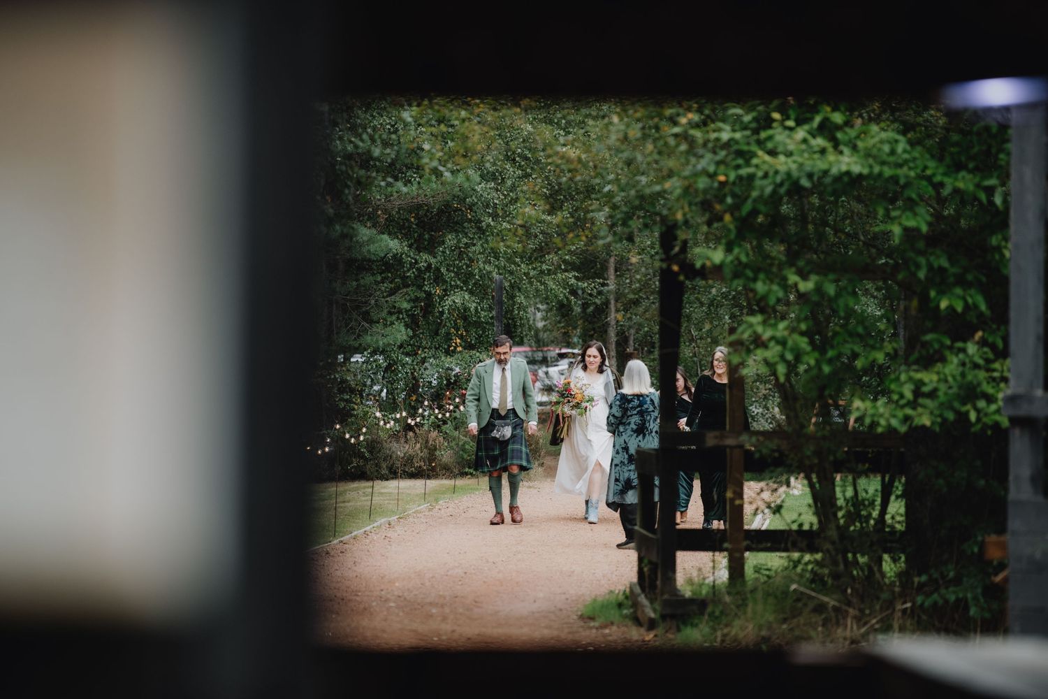 Bride and groom in Scottish tartan kilt walking through a wooden gate in a forest setting.