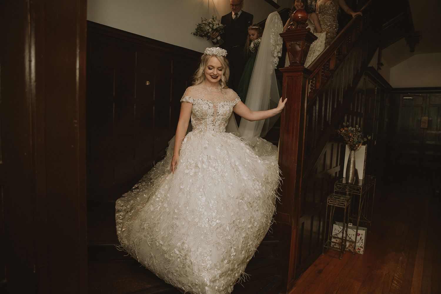 Bride in an elegant off-shoulder ball gown with feather details descends a wooden staircase in a historic venue.