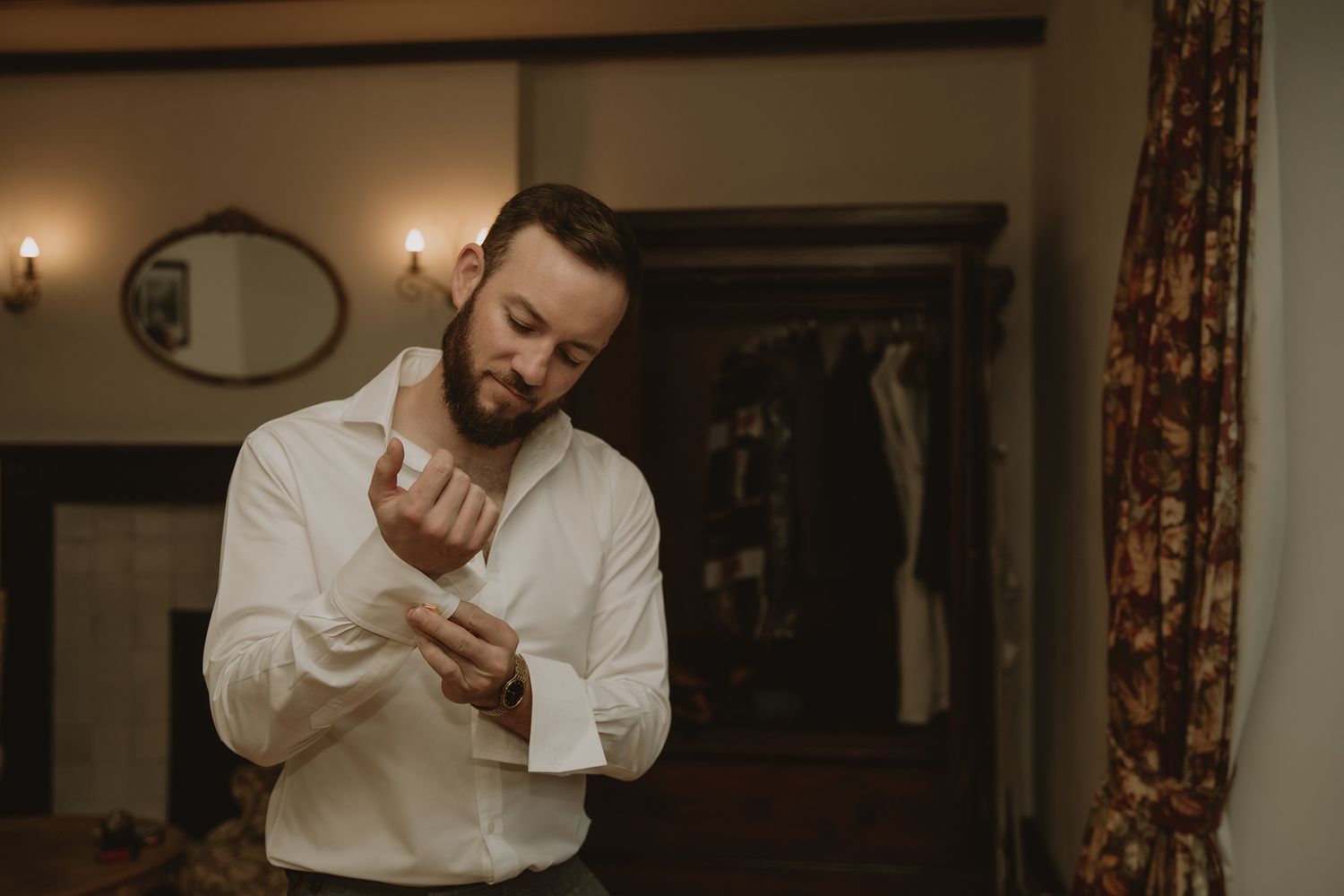 A groom in a white dress shirt fastening his cufflinks while getting ready in an elegant room.