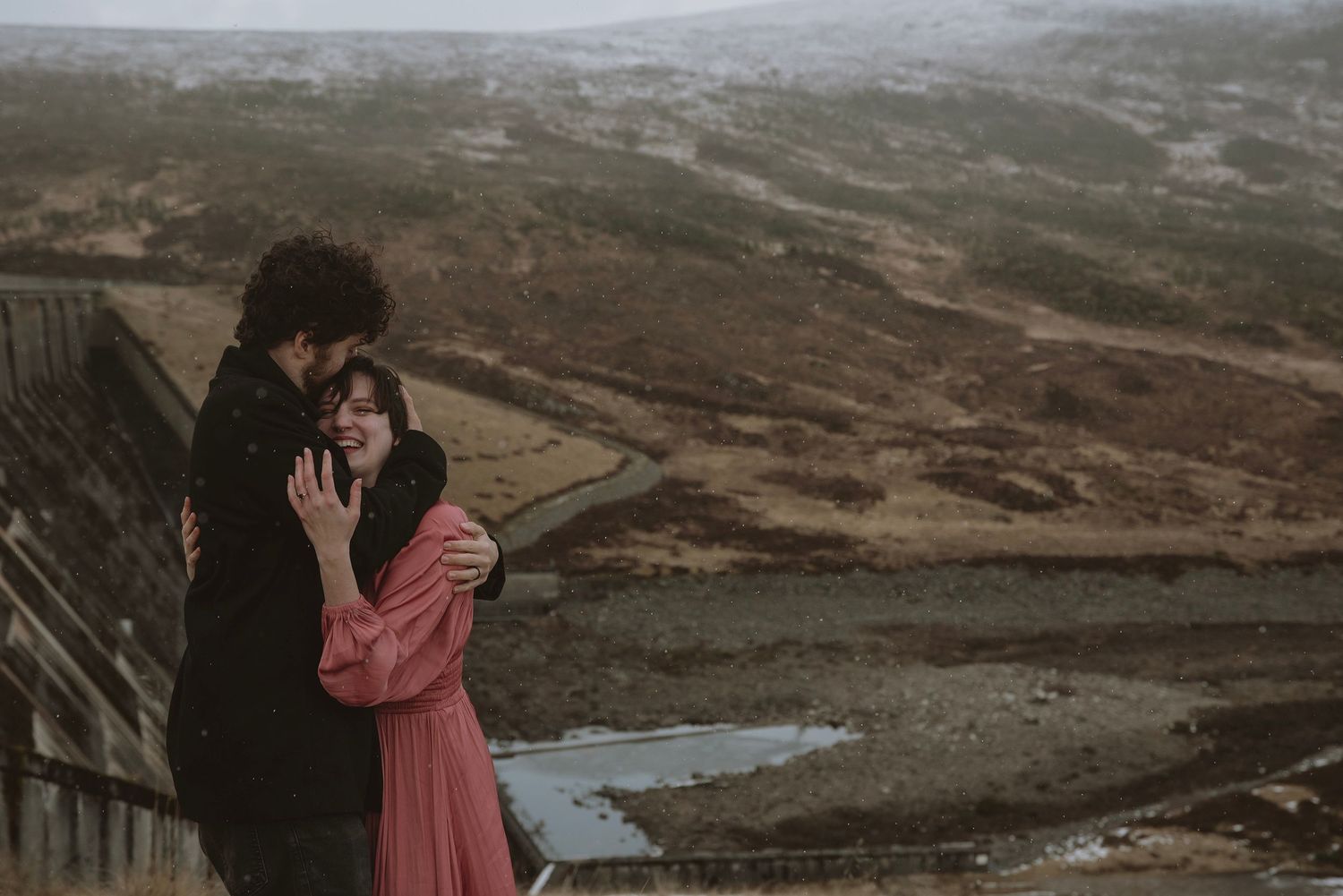 Couple embracing romantically on a misty hilltop overlooking a scenic valley and reservoir in moody weather.
