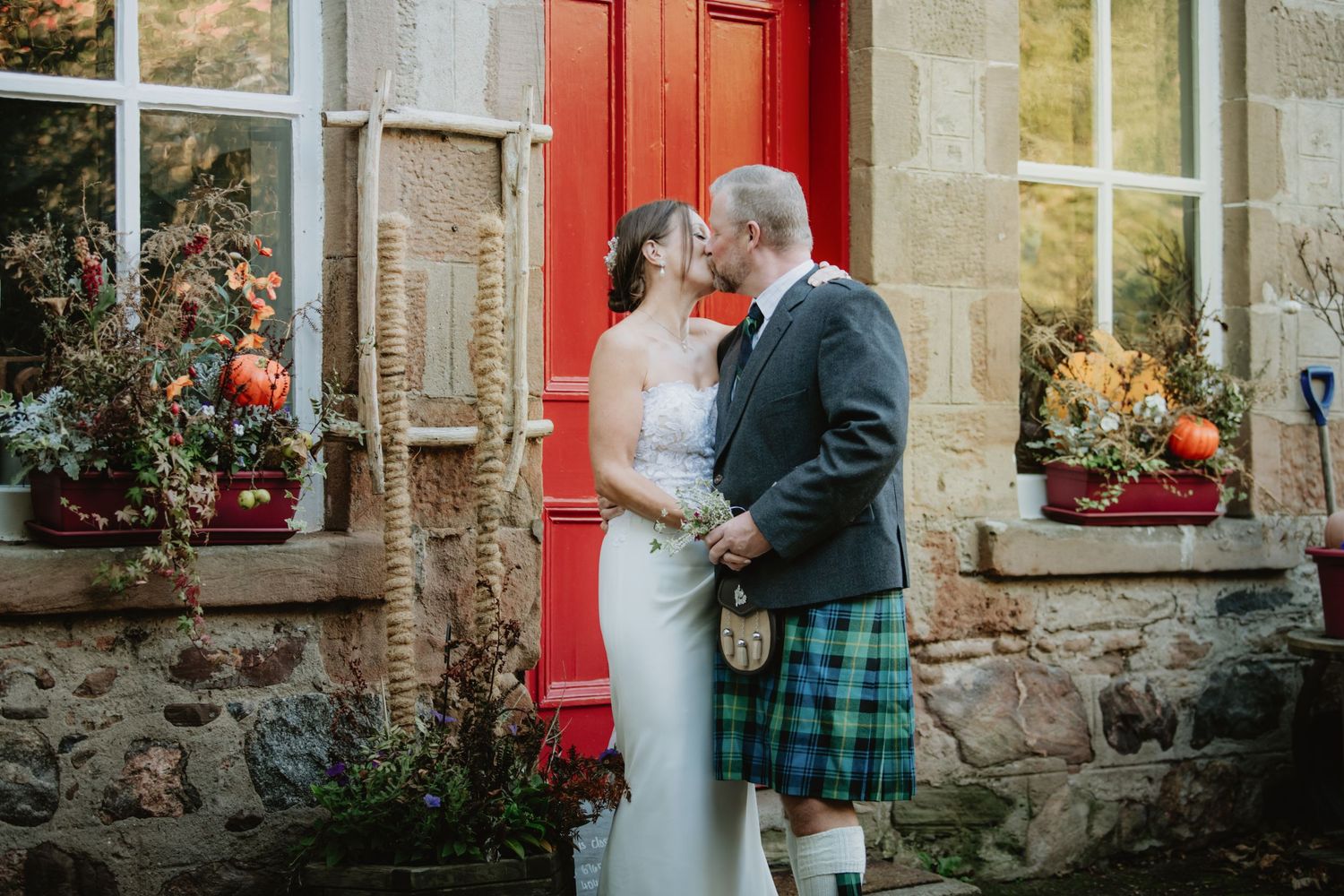 Bride and groom share a romantic kiss outside a rustic stone building with a red door.