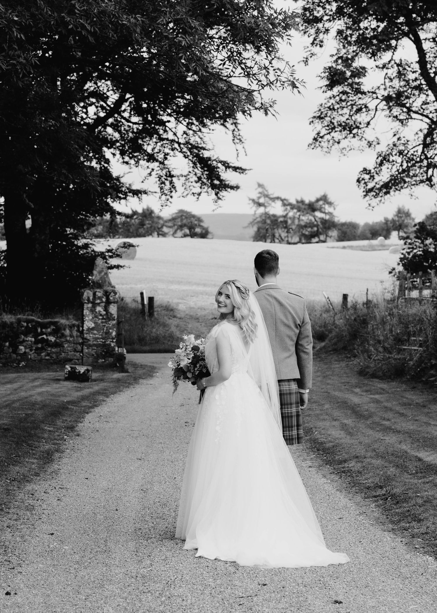 Bride and groom in Scottish kilt walk along gravel path surrounded by countryside in black and white.