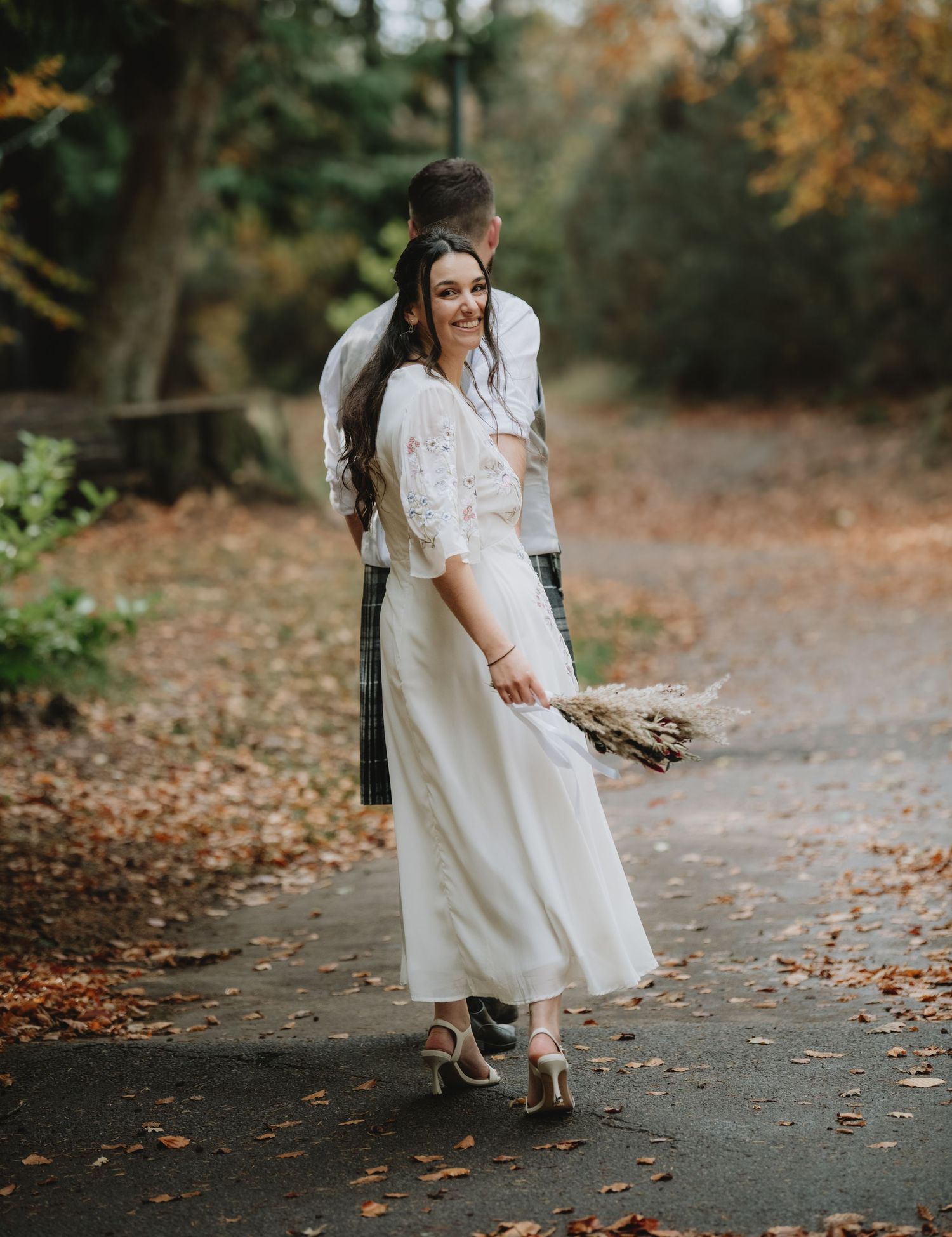 Bride in floral lace dress holding dried bouquet while groom kisses her head on autumn woodland path.