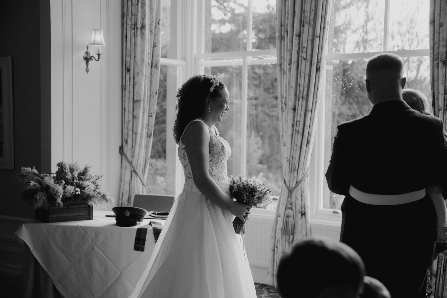 Bride in lace wedding gown stands beside groom in military uniform during intimate wedding ceremony by window.