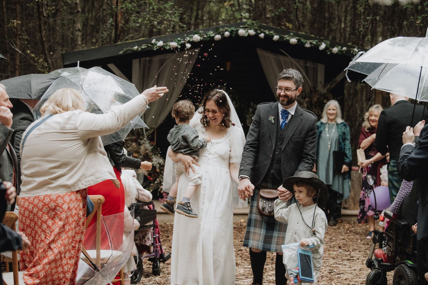 Newlyweds walk through confetti shower with guests celebrating at woodland wedding.