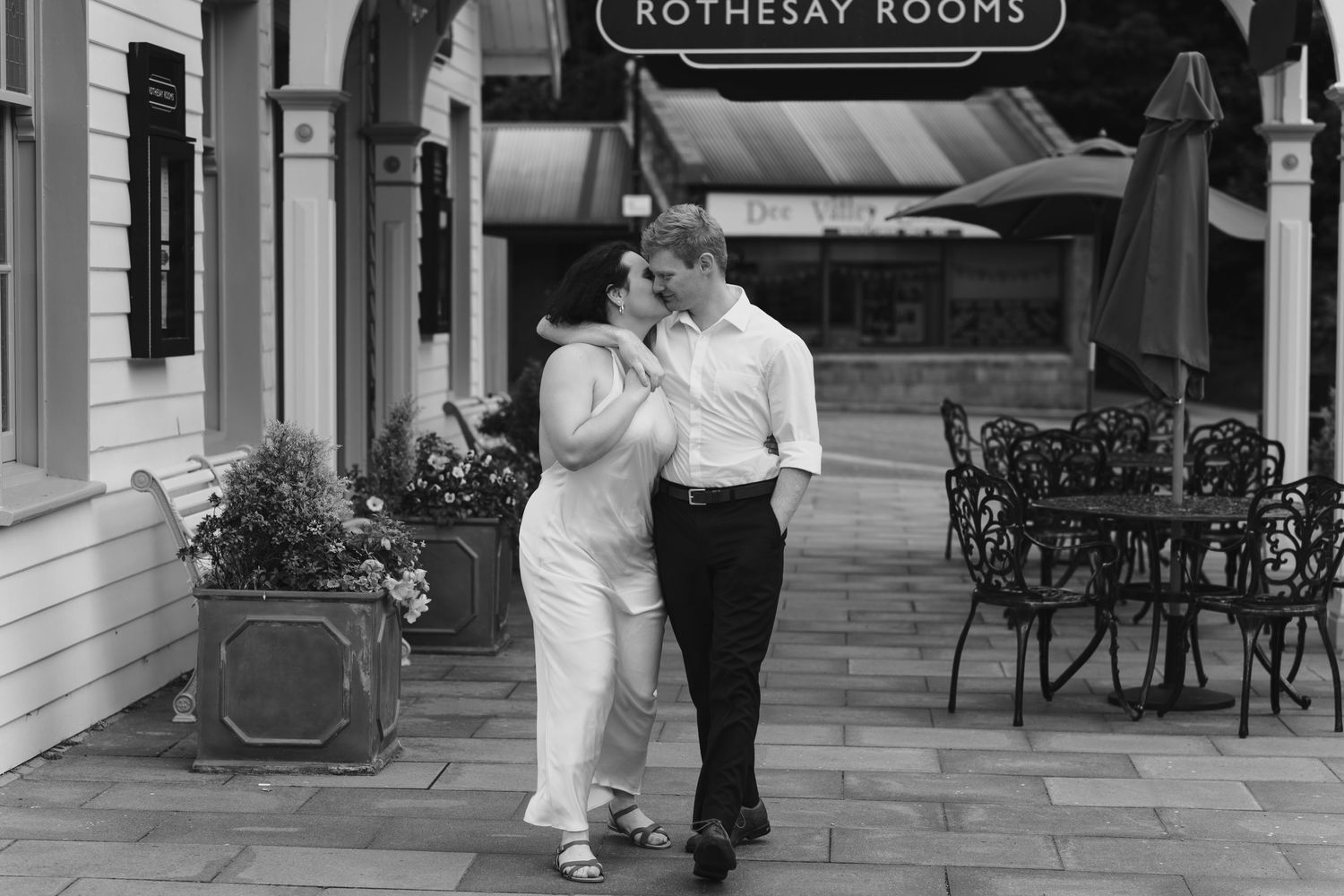 Couple embracing and smiling outside the Rothesay Rooms venue with iron patio furniture nearby.