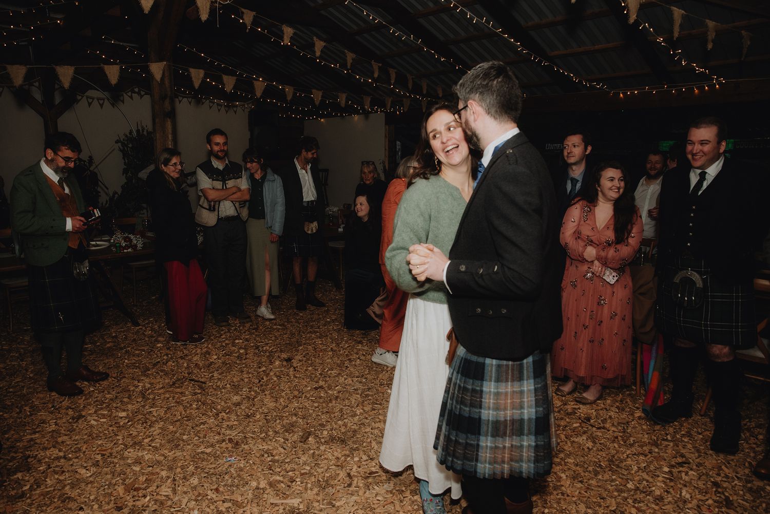 Bride and groom sharing their first dance in a rustic barn venue with string lights and wedding guests watching.