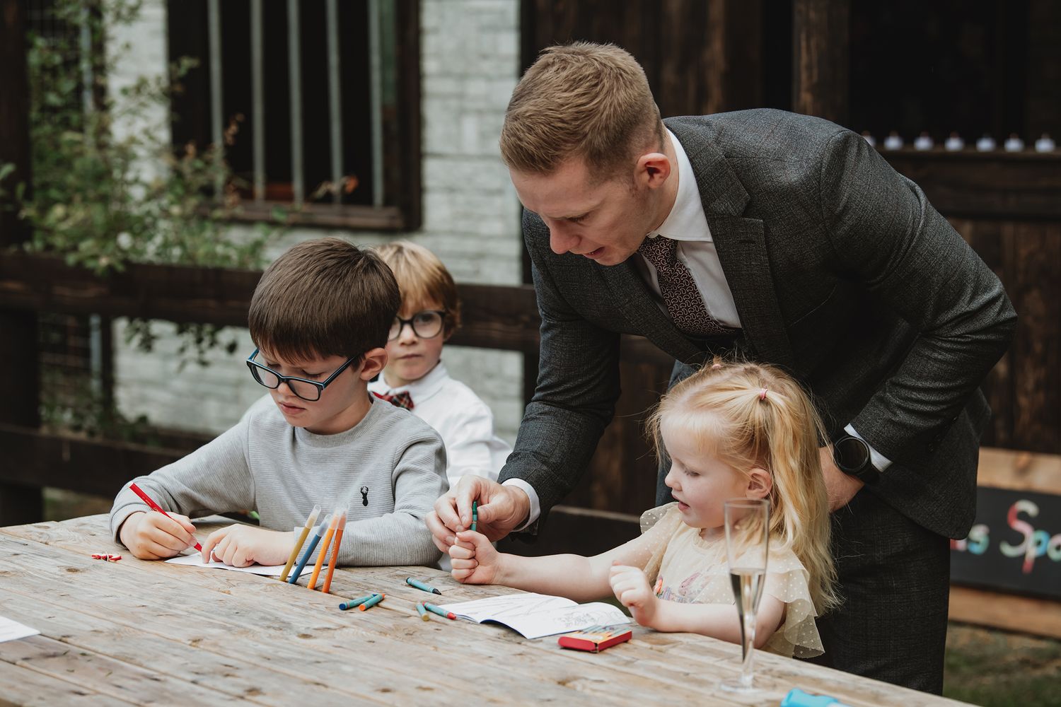 A man in a suit helps children with a drawing activity at an outdoor wooden table.