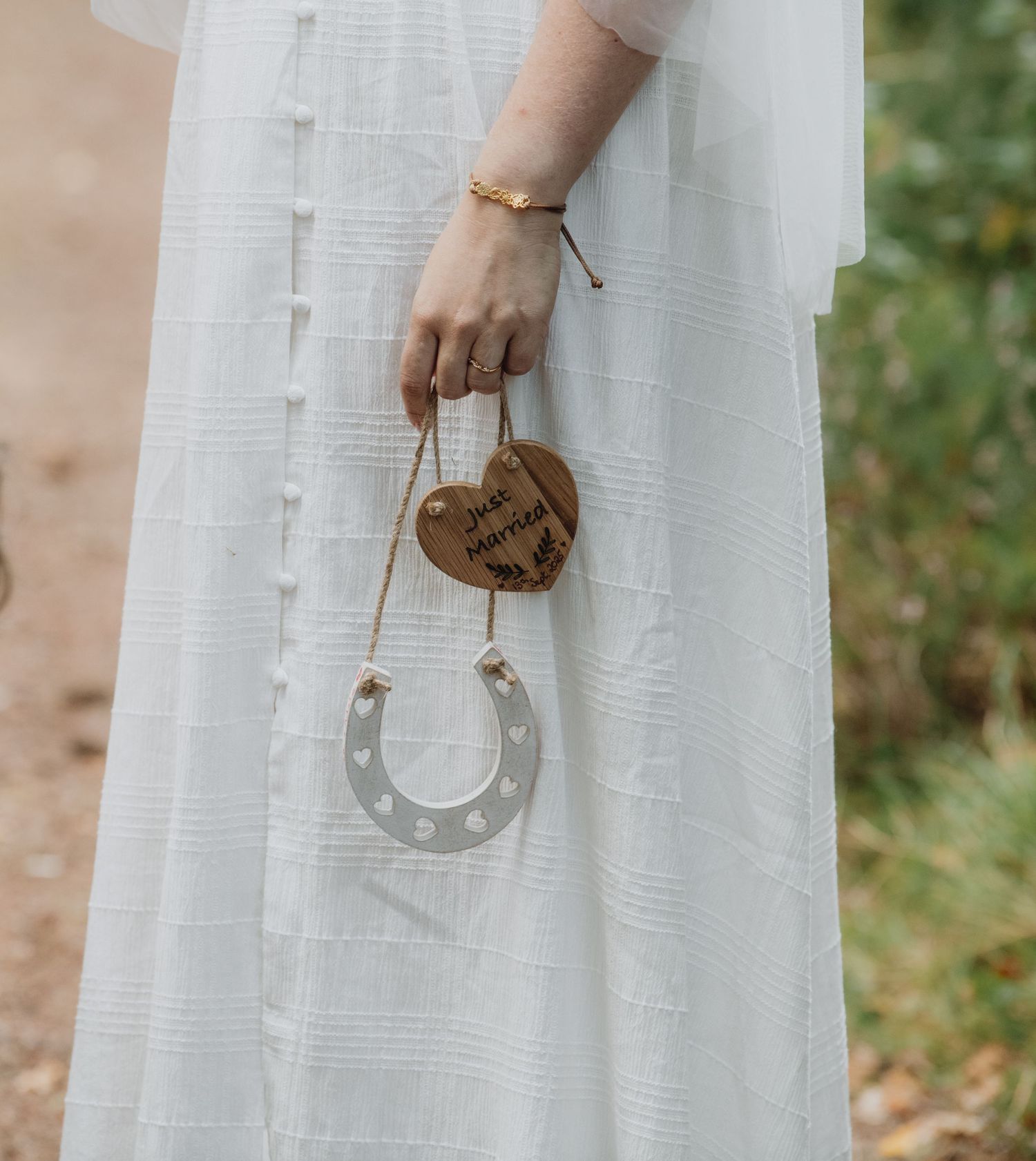 Bride holds lucky horseshoe charm with wooden heart tag at outdoor woodland wedding ceremony.