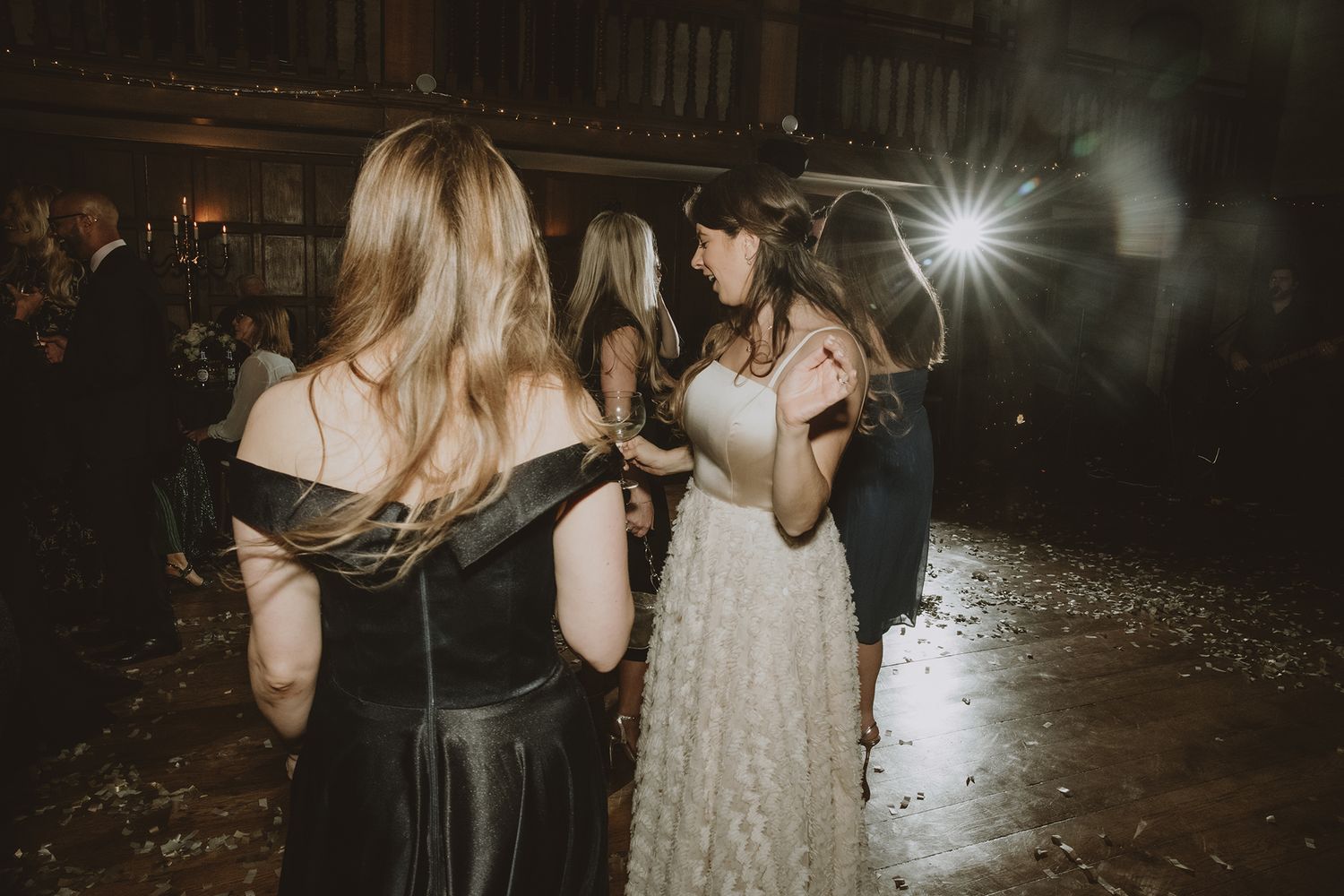 Bride dancing with guests at elegant wedding reception with dramatic lighting and confetti on the floor.